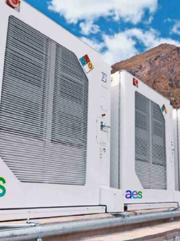 A person wearing a hard hat and safety vest walks past large white industrial units with 'aes' logos, set against a mountainous backdrop under a blue sky with clouds.