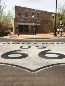 A street in Winslow, Arizona, featuring a large Route 66 emblem painted on the road. In the background, there is a brick building with a sign that reads 'Winslow Arizona.' Trees and street lamps line the sidewalk, and a red truck is parked nearby.