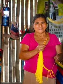 A woman in a pink shirt stands in a wooden room, weaving a yellow thread into a hammock. A calendar and a lantern hang on the wall behind her.