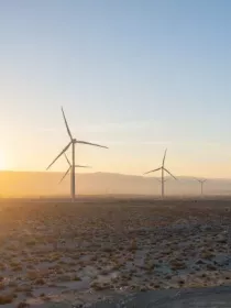 A landscape with multiple wind turbines on a desert plain at sunrise. The sun is partially visible on the horizon, casting a warm glow over the turbines and the rocky terrain.