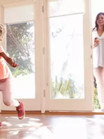 A young child joyfully runs across a sunlit room with hardwood floors. In the background, a smiling family, including an adult holding a baby, enters through a glass door. Bright natural light streams in, creating a warm atmosphere.