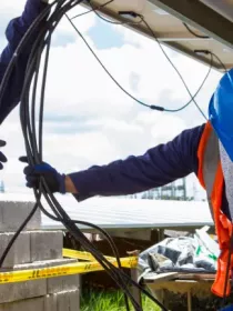 Two workers wearing blue helmets and safety gear are installing cables under a solar panel. One worker is holding the cables while the other is securing them. The background shows more solar panels and a clear sky.