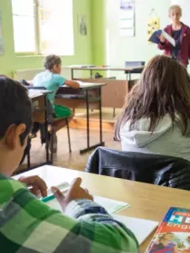 A classroom with students seated at desks, writing in notebooks. A teacher stands at the front near a whiteboard, holding a book. The classroom walls are light green, and a textbook titled 'Hello! English for the 6th grade' is visible on a desk in the foreground.