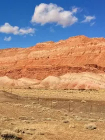 A landscape featuring red rock formations under a clear blue sky with a few clouds. The foreground shows dry grass and sparse vegetation, with a fence running parallel to the rock formations.