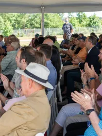 A diverse group of people seated under a tent, attentively watching an event and clapping. The setting appears to be outdoors with trees visible in the background.