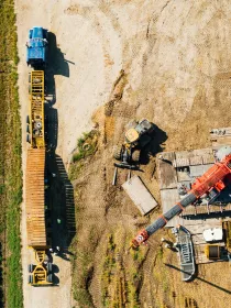 Aerial view of a construction site with a blue truck and a yellow trailer parked on a dirt path next to a green field. Nearby, a construction vehicle is working on a dirt area with wooden planks and a red crane. Several people are visible around the site.