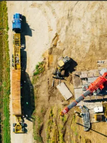 Aerial view of a construction site next to a green field. The site contains a large red crane, several wooden pallets, and construction vehicles, including a blue truck and a yellow excavator. The ground is a mix of dirt and grass.