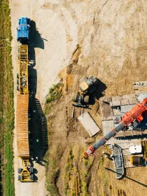 Aerial view of a construction site with a blue truck and a yellow trailer on a dirt path next to a green field. A red crane and construction materials are on the right side.