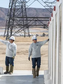 Two workers wearing hard hats and safety gear inspect large white industrial units outdoors. A metal tower and dry landscape are visible in the background.
