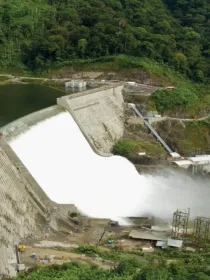 Aerial view of a large dam with water flowing over its spillway, surrounded by lush green forested hills.