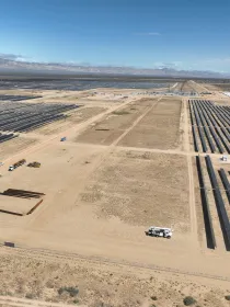 Aerial view of a large solar farm in a desert landscape. Numerous rows of solar panels are arranged in parallel lines across the sandy terrain. Several vehicles and construction equipment are visible near the center of the image. Mountains are seen in the distance under a clear blue sky.