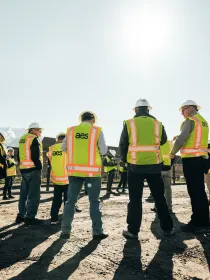 A group of people wearing yellow safety vests and hard hats stand outside on a sunny day. They are gathered on a dirt surface with mountains visible in the background.