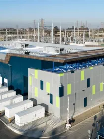Aerial view of a large industrial building surrounded by electrical equipment and power lines. The building has a modern design with blue and gray panels and green accents. Rows of white containers are aligned along one side. The surrounding area includes roads and additional industrial structures under a clear blue sky.