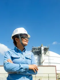 A person wearing a hard hat and safety glasses stands with arms crossed in front of a large industrial tank under a clear blue sky.
