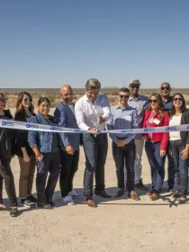 A group of people stand outdoors on a sunny day, participating in a ribbon-cutting ceremony. A man in the center is cutting a ribbon with large scissors. The ribbon has the AES logo on it. The group is smiling and appears to be celebrating an event. The background shows a clear sky and open landscape.