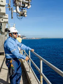 A worker wearing a blue uniform and white hard hat stands on a platform overlooking a large body of water. The worker is holding onto a metal railing. In the background, there is a small boat on the water and a distant shoreline with industrial structures.