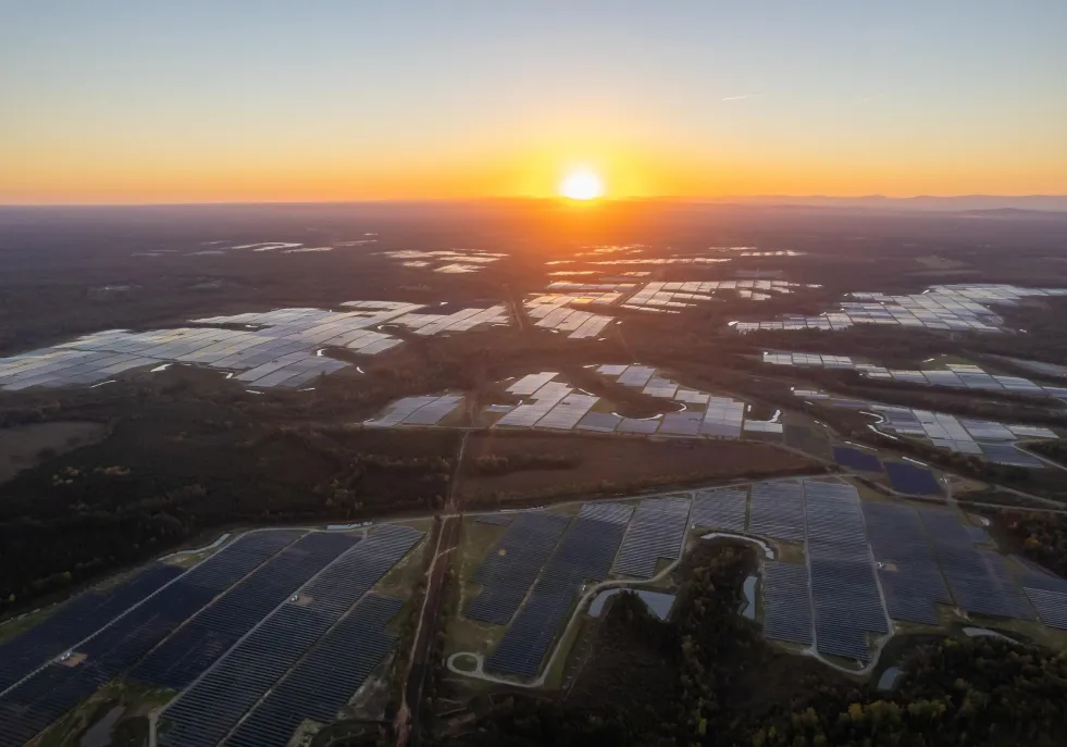 Aerial view of a vast solar farm at sunset, with rows of solar panels reflecting the orange and yellow hues of the sky.
