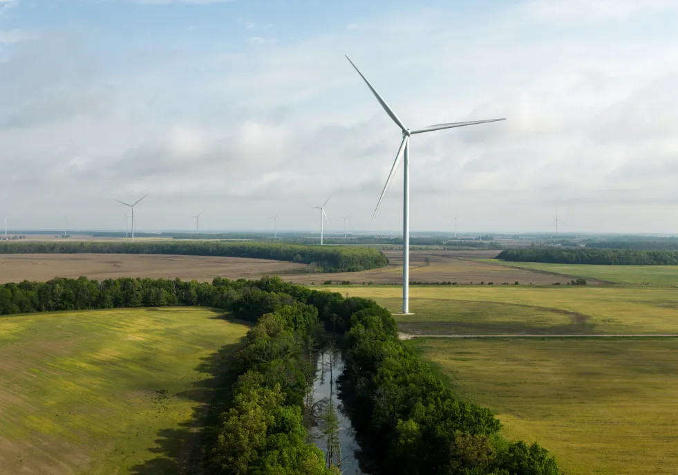 Aerial view of a landscape with multiple wind turbines on a green field under a cloudy sky. A narrow river runs through the foreground surrounded by trees.