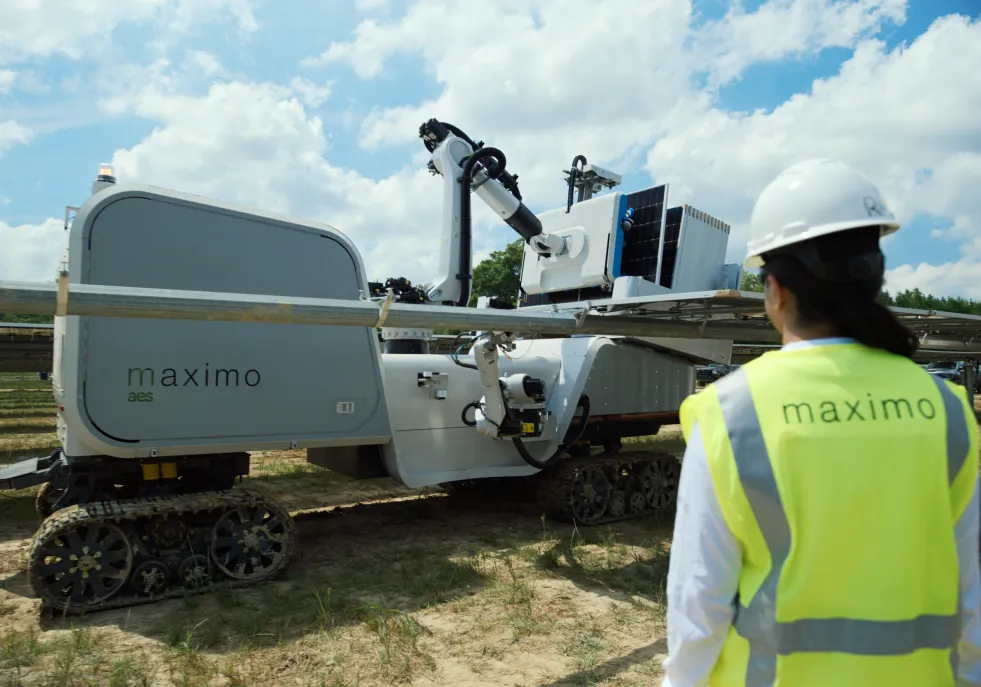 A person in a safety vest and hard hat stands near a Maximo AES robotic machine on tracks in a field under a cloudy sky.