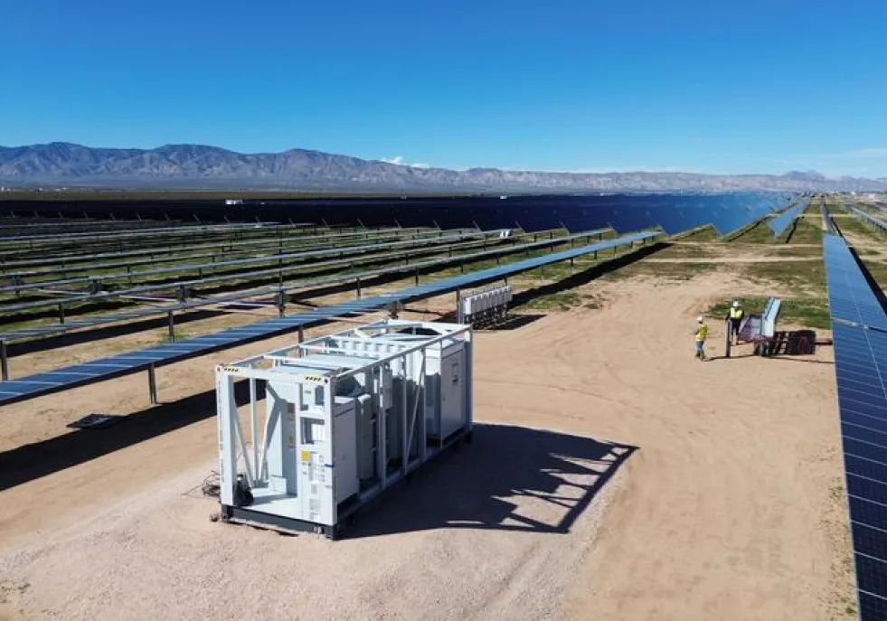A large solar power facility with rows of solar panels stretching into the distance. A white utility box is in the foreground on a dirt path. Mountains are visible in the background under a clear blue sky.