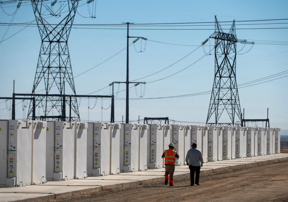 Two people walk along a row of large white battery storage units under high-voltage power lines against a clear blue sky.