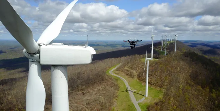Aerial view of a wind farm with several wind turbines on a hilly landscape. A drone is flying near one of the turbines under a cloudy sky.