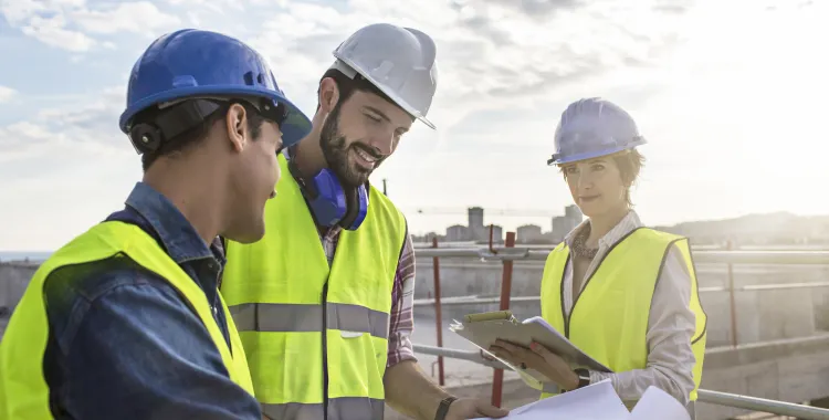 Three construction workers wearing safety vests and helmets stand on a rooftop. Two men are looking at a large blueprint, while a woman holds a clipboard, smiling. The background shows a cityscape under a partly cloudy sky.