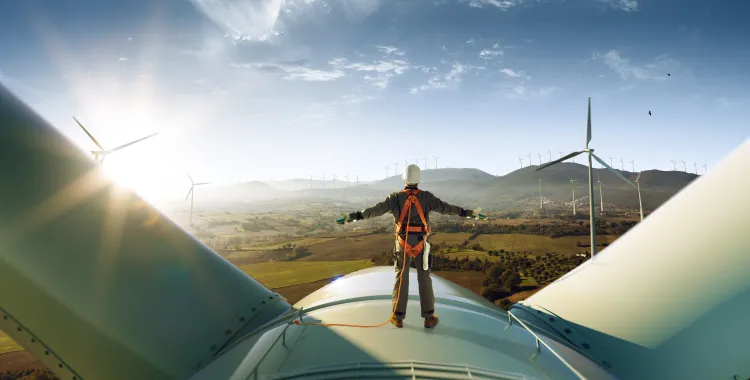 A person in safety gear stands on top of a wind turbine, arms outstretched, overlooking a landscape with multiple wind turbines, fields, and distant hills under a clear blue sky.