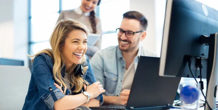 Three colleagues in a bright office smiling and looking at a computer screen. A woman in the foreground is laughing, while a man beside her and another woman standing behind them also smile.