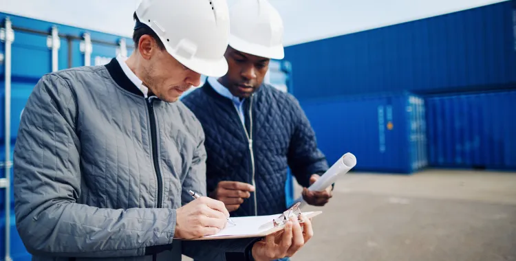 Two construction workers wearing white helmets review documents on a clipboard at a shipping container yard.