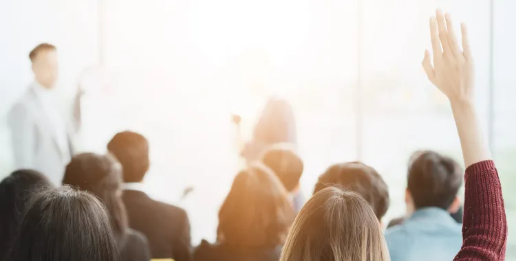 A group of people attending a seminar, with one person raising their hand to ask a question. Two speakers are visible in front of a bright window.