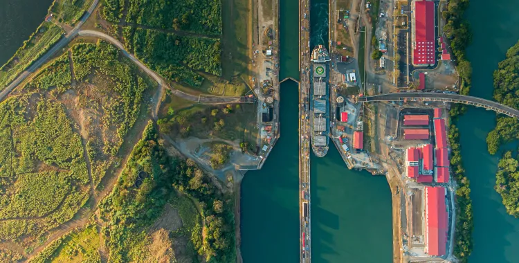 Aerial view of a canal with a ship passing through a lock. The canal is surrounded by lush green vegetation on the left and industrial buildings with red roofs on the right. The lock gates are open, allowing the ship to move through. Roads and pathways are visible around the canal area.