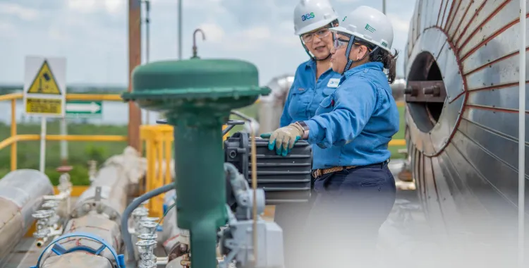 Two workers in blue uniforms and hard hats inspect equipment at an industrial site. One holds a tool while the other observes. Pipes and machinery surround them under a cloudy sky.