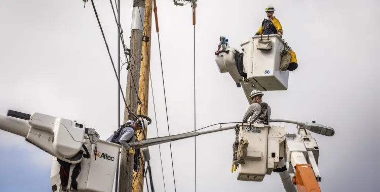 Three utility workers in bucket trucks repair electrical lines on a power pole under a cloudy sky.