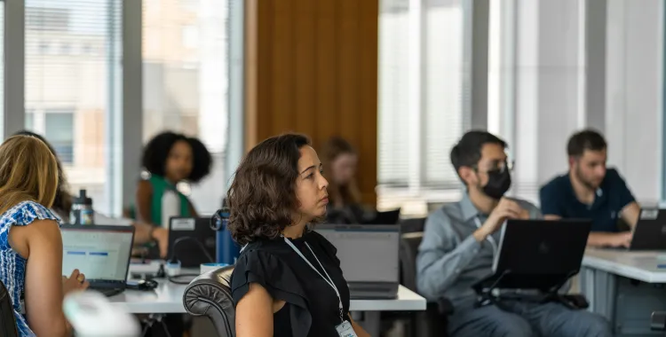 A group of adults sitting at desks in a modern office or classroom, focused on a presentation. Some people are using laptops, and one person is wearing a face mask. Large windows let in natural light.