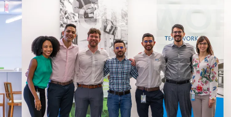 Seven diverse colleagues stand together smiling in an office environment, showing teamwork and camaraderie. The background features a motivational wall with the word 'TEAMWORK' partially visible.
