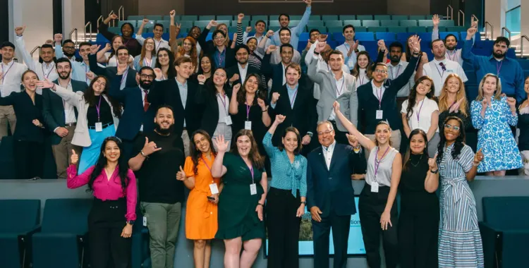 A large, diverse group of people in business and casual attire pose together in an auditorium, smiling, raising their hands, and making celebratory gestures. The setting suggests a professional or academic event with an upbeat and inclusive atmosphere.