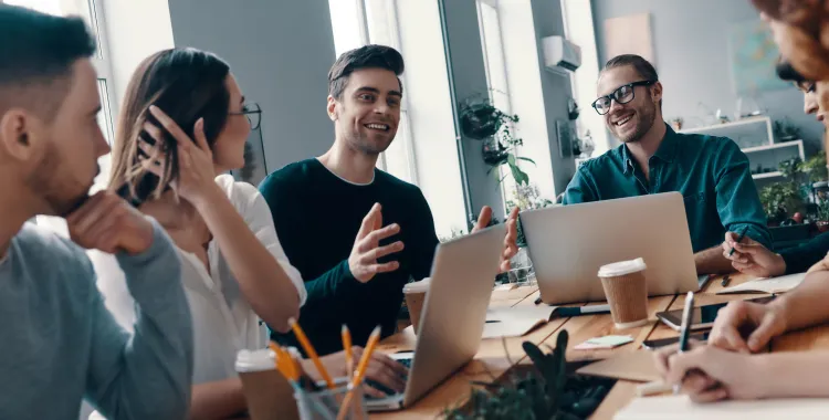 A group of young professionals having a discussion around a wooden table in a modern office, with laptops, notebooks, and coffee cups in front of them. They are smiling and engaged in conversation, with natural light coming through large windows.