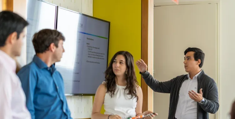 Four people in business attire are engaged in a discussion during a meeting. One man is presenting in front of a large screen displaying a presentation, while the others listen attentively in a modern office setting.