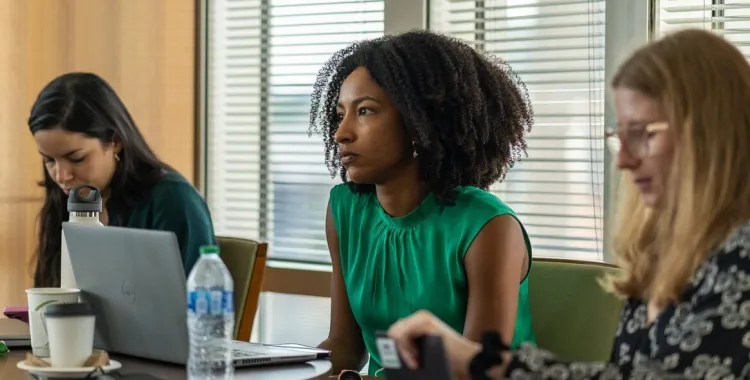 Three women sit at a conference table with laptops, notebooks, and water bottles, focused on a meeting in a modern office with large windows and blinds.