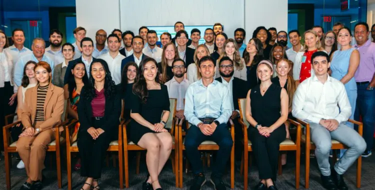 A large group of professionally dressed people pose together indoors, seated and standing in rows, smiling at the camera in an office setting.