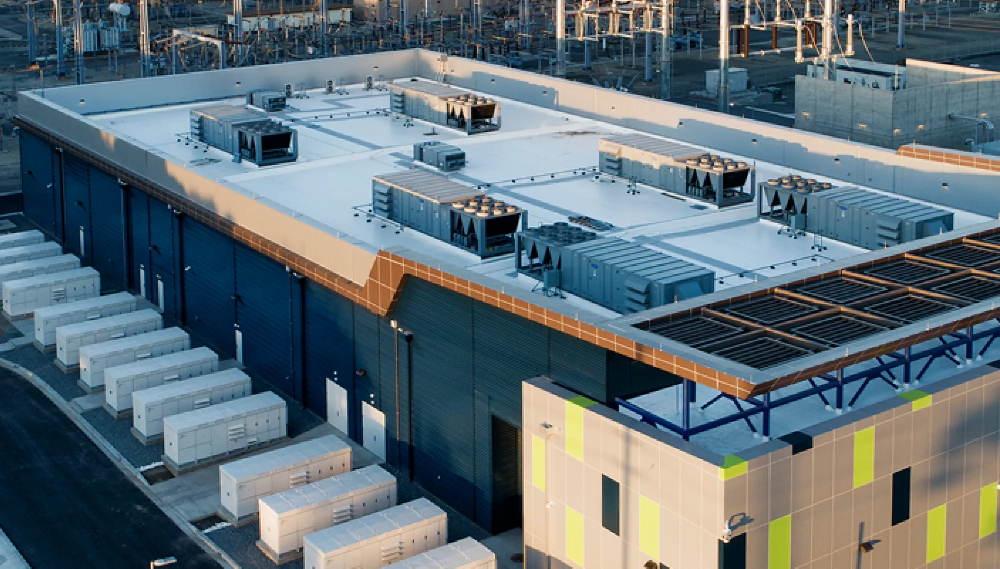 Aerial view of a modern industrial data center with a flat roof, multiple cooling units, and rows of white storage containers on the ground. The building is surrounded by power lines and other industrial structures.