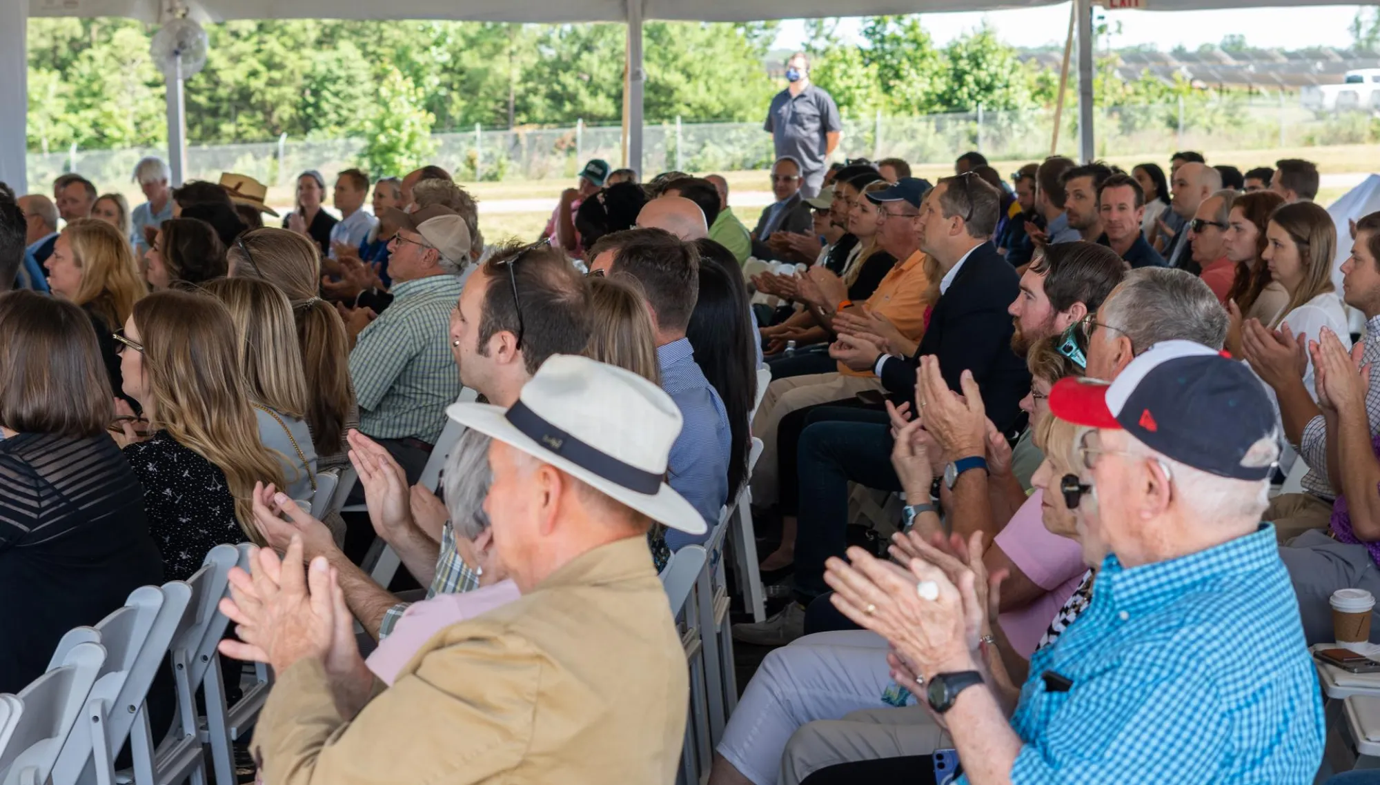 A large group of people seated under a tent, clapping and facing forward, with trees visible in the background.