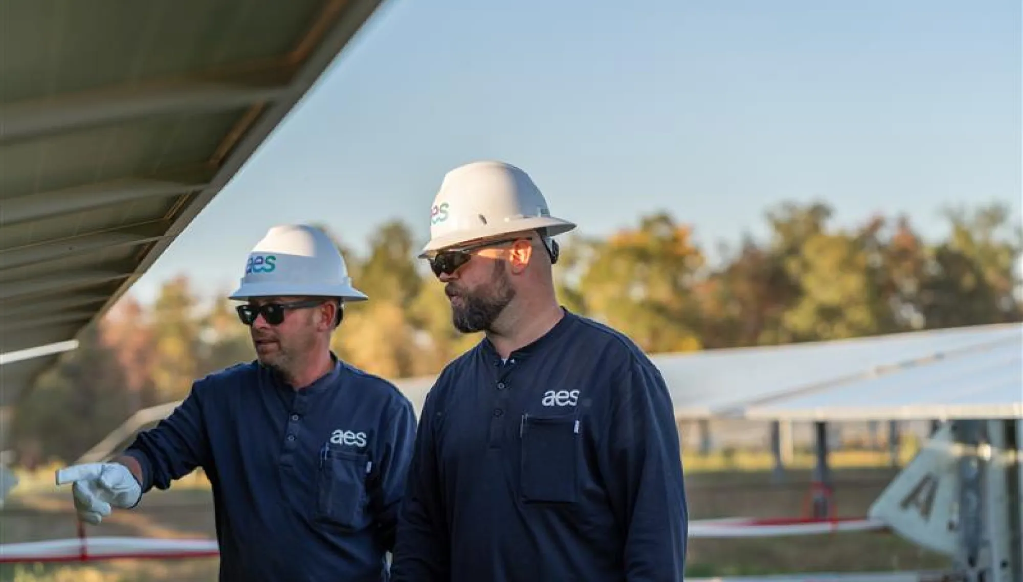 Two workers wearing hard hats and sunglasses inspect solar panels outdoors. They are dressed in dark uniforms with a logo on the chest. The background shows a field with more solar panels and trees under a clear sky.