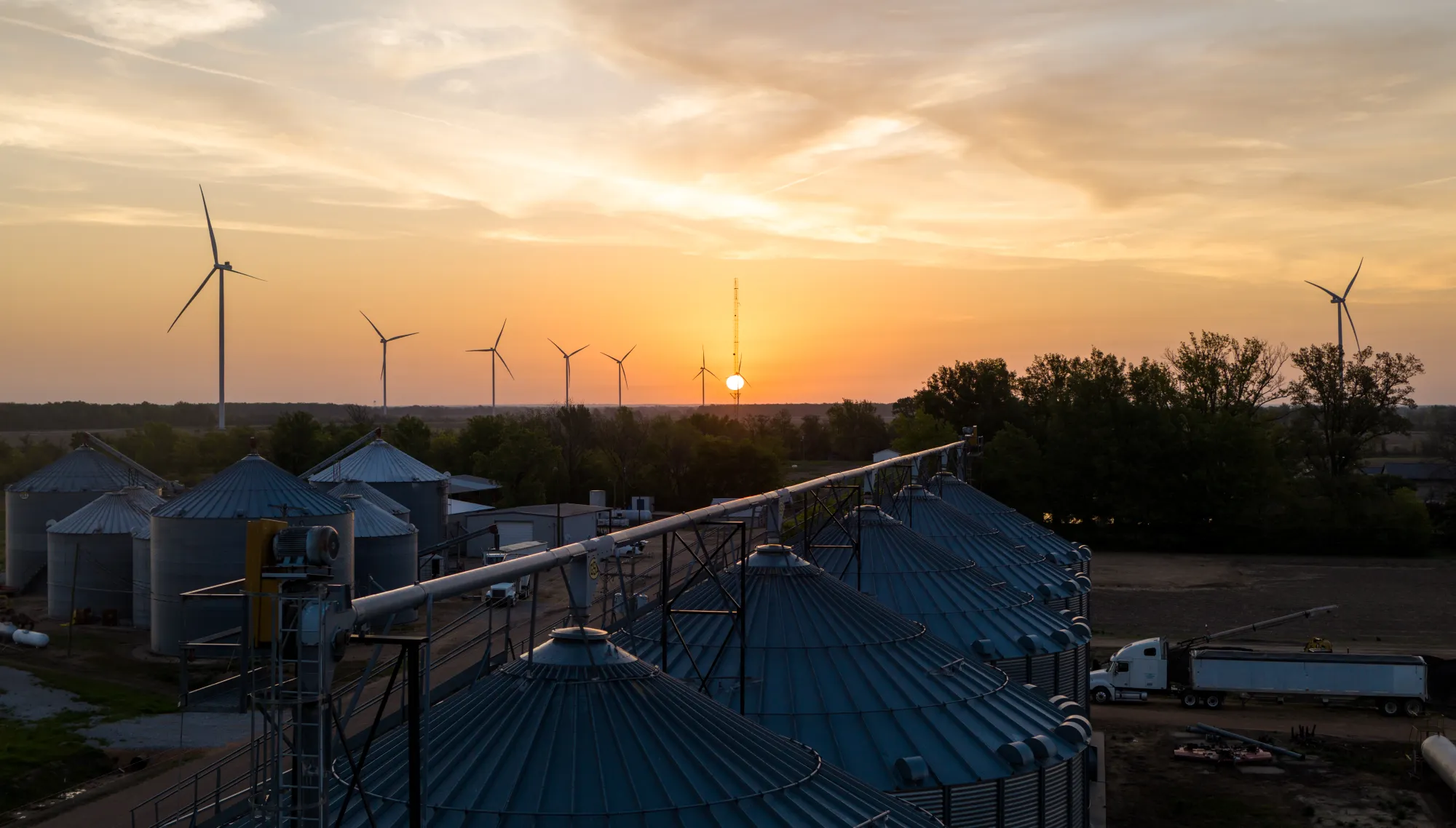 A rural landscape at sunset featuring several wind turbines and large metal silos. The sky is filled with orange and yellow hues as the sun sets on the horizon. Trees and fields surround the silos, and a truck is parked nearby.