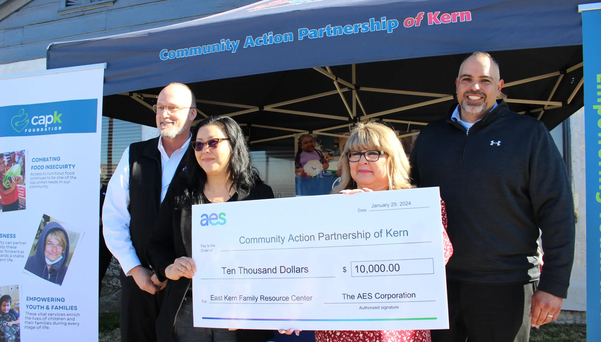 Four people stand in front of a tent labeled 'Community Action Partnership of Kern.' They are holding a large ceremonial check for $10,000 from AES Corporation to the Community Action Partnership of Kern, dated January 29, 2024. A banner and informational posters about food insecurity and community support are visible in the background.