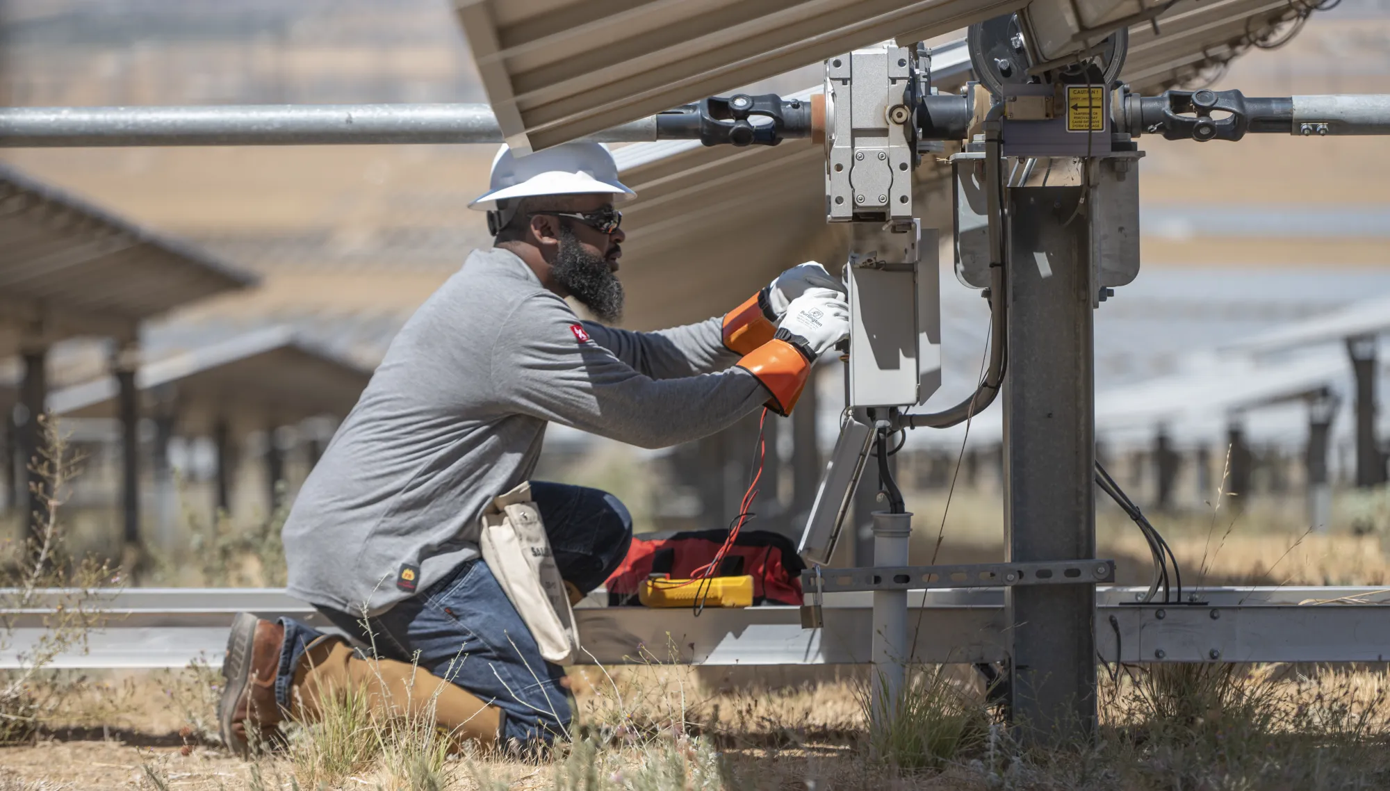 A technician wearing a hard hat and gloves kneels under a solar panel, working on electrical equipment. The technician is using tools and has a tool bag nearby. Rows of solar panels are visible in the background, set in a dry, grassy field.