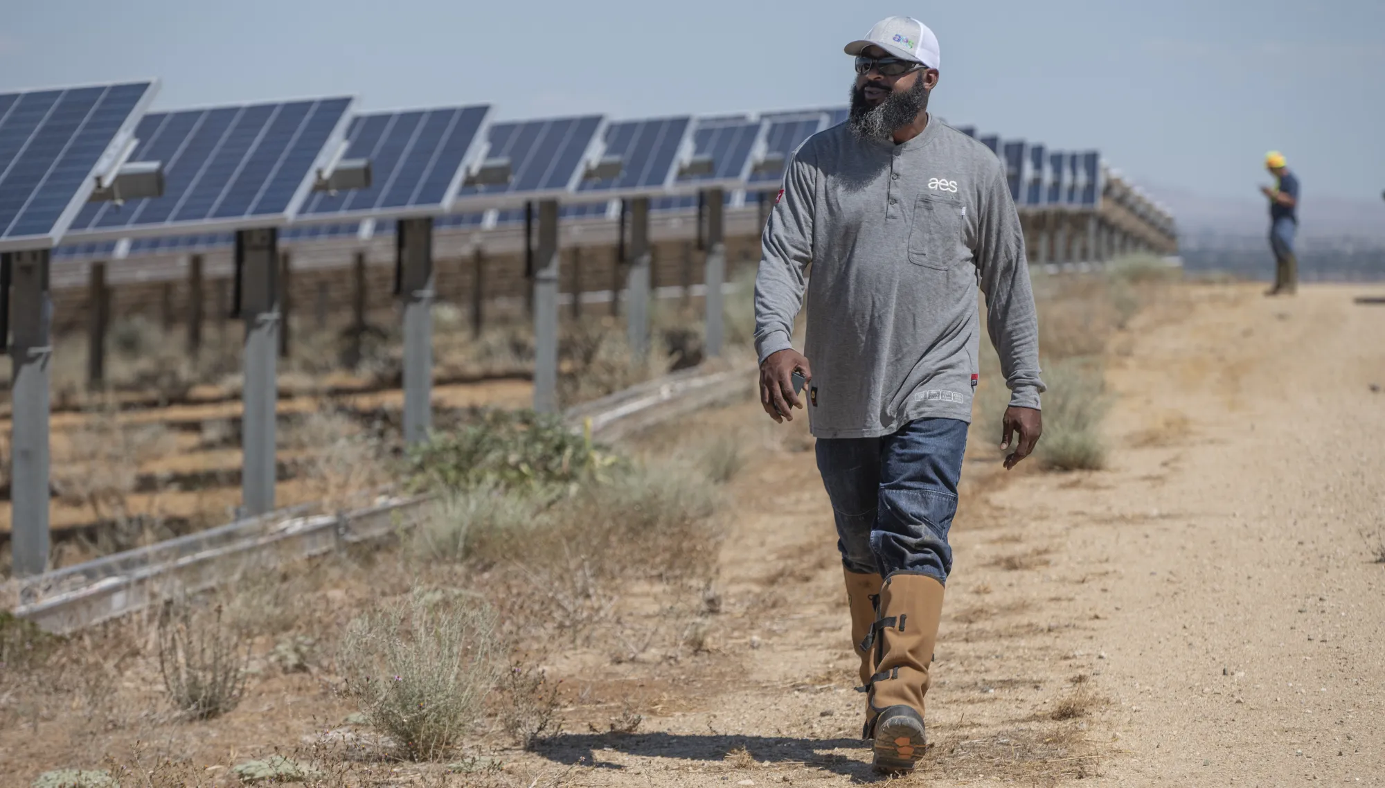 A person wearing a gray shirt with an AES logo, jeans, and boots walks along a dirt path beside rows of solar panels. Another person in the background is wearing a hard hat and looking at a device.