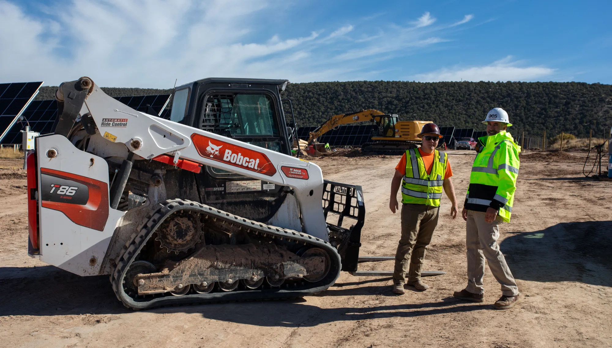 Two construction workers in safety vests and helmets stand beside a Bobcat T86 compact track loader on a dirt construction site. Solar panels and another excavator are visible in the background under a clear blue sky.