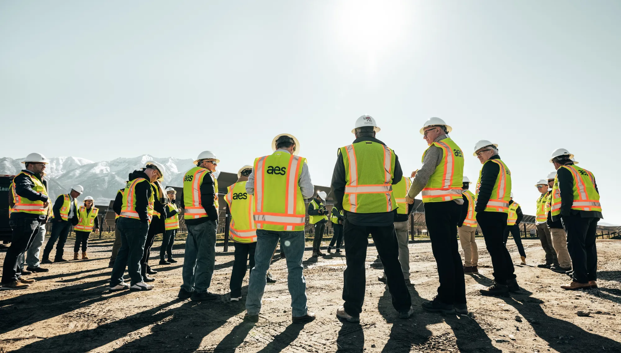 A group of people wearing yellow safety vests and hard hats stand outside on a sunny day. They are gathered on a dirt surface with mountains visible in the background.